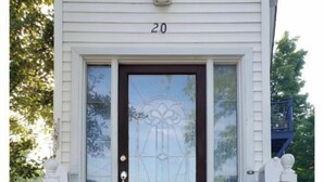 A white house with the number 20 on the front door, a glass door with a decorative pattern, a small porch with steps, and two potted plants with pink flowers. - Fairport Village Triangle Home on the Erie Canal (Fairport)