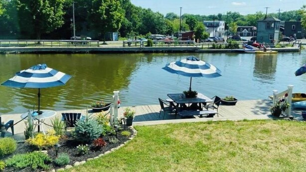 A waterfront patio with two umbrellas, a table, and chairs. - Fairport Village Triangle Home on the Erie Canal (Fairport)