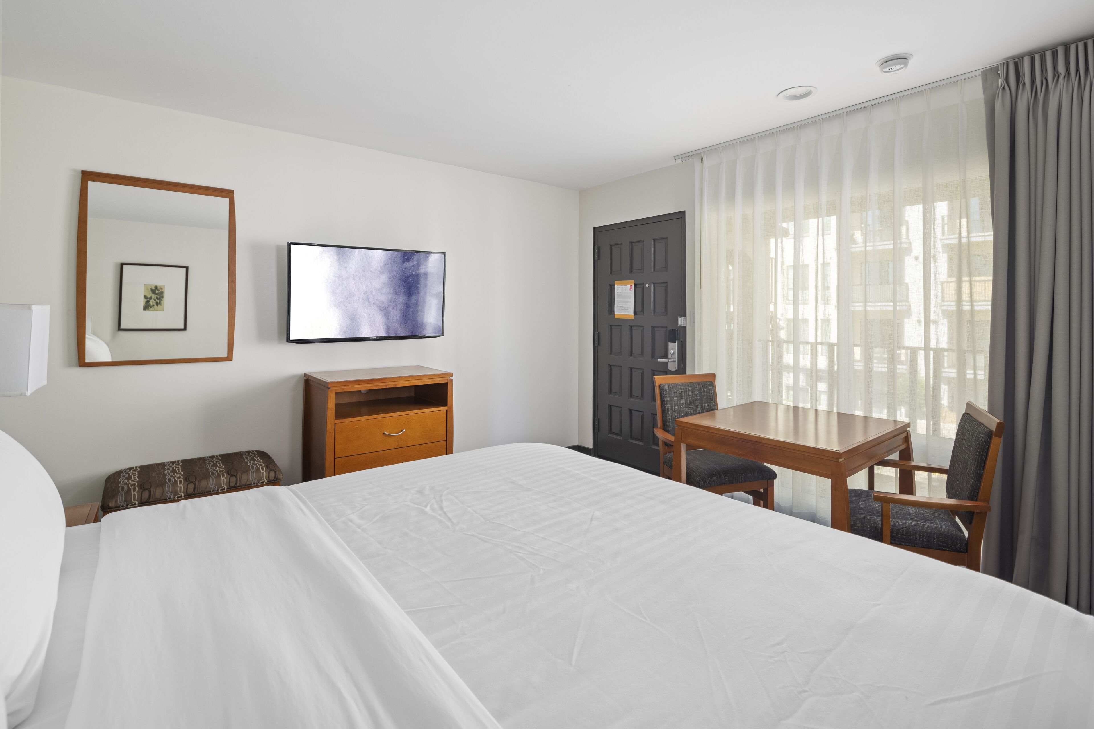 A hotel room with a large bed, a wooden desk with two chairs, a television, and a door with a notice.