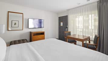 A hotel room with a large bed, a wooden desk with two chairs, a television, and a door with a notice.