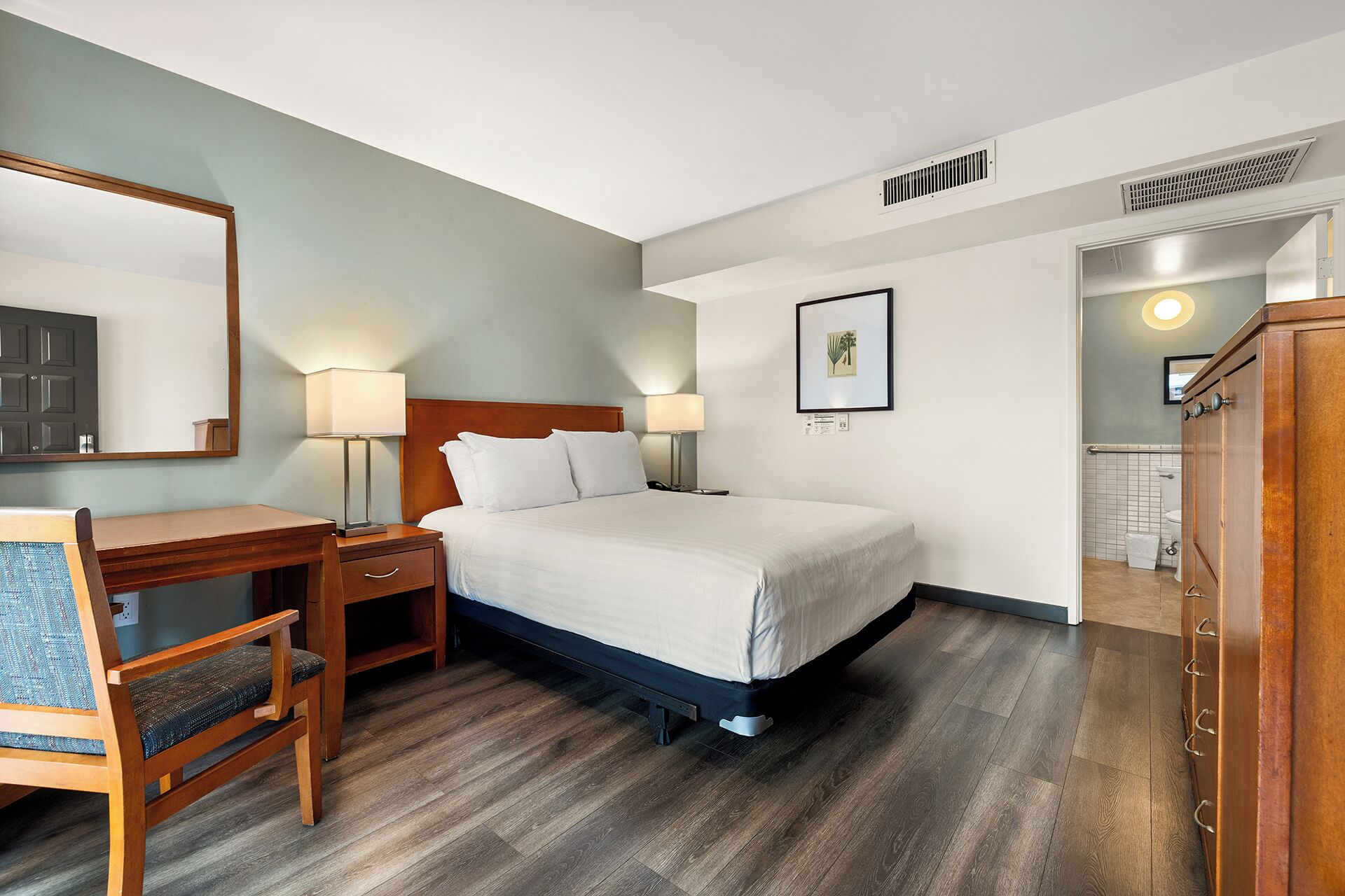A hotel room with a bed, bedside tables, a desk, a chair, a mirror, a framed picture, and a door leading to a bathroom.