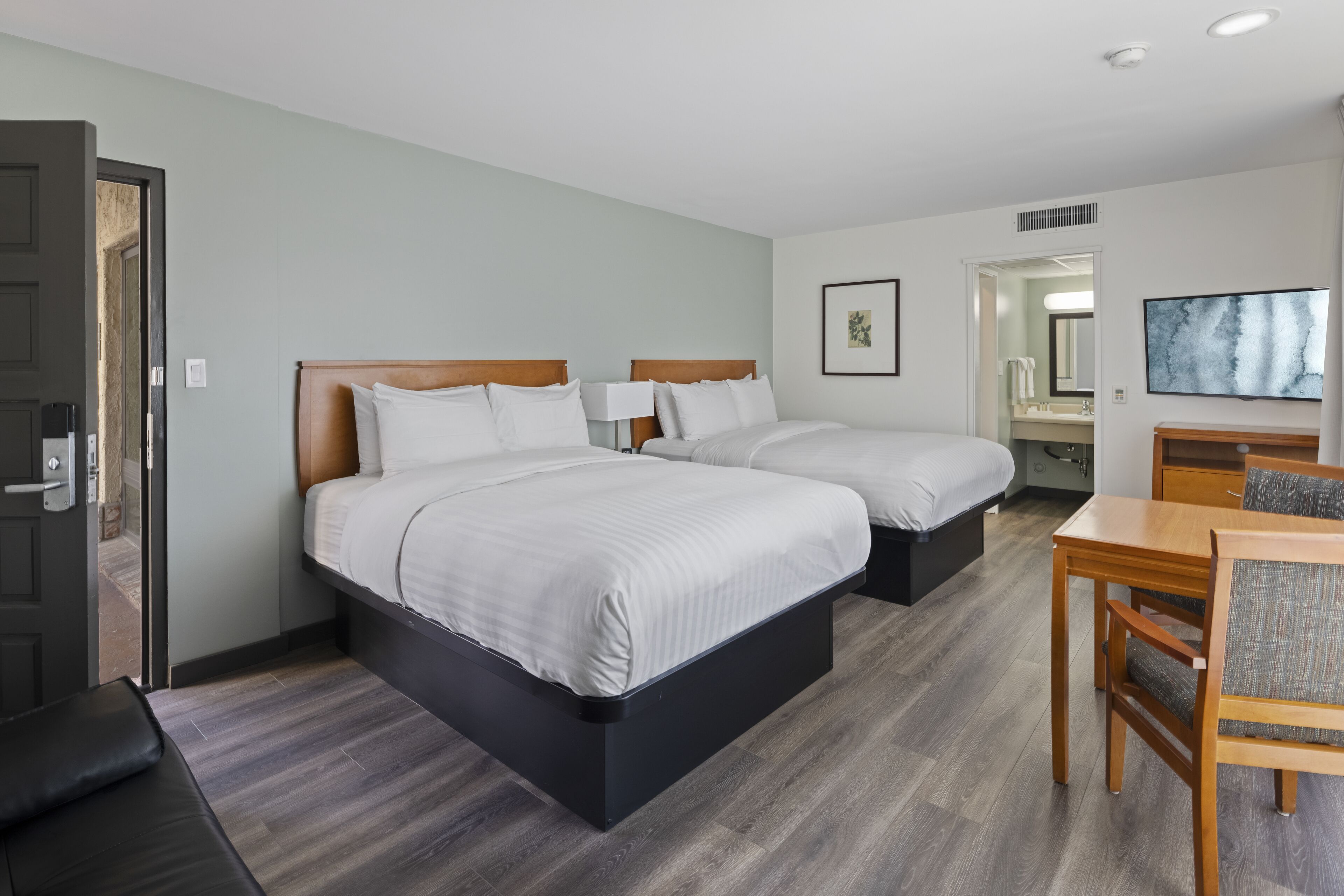 A hotel room with two beds, a desk, a chair, a TV, and a bathroom visible through an open door.