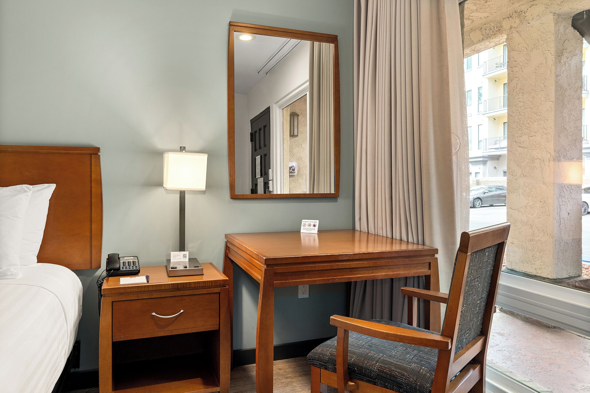 Una habitación de hotel con una cama, un escritorio con lámpara, una silla, un espejo y una ventana con cortinas.