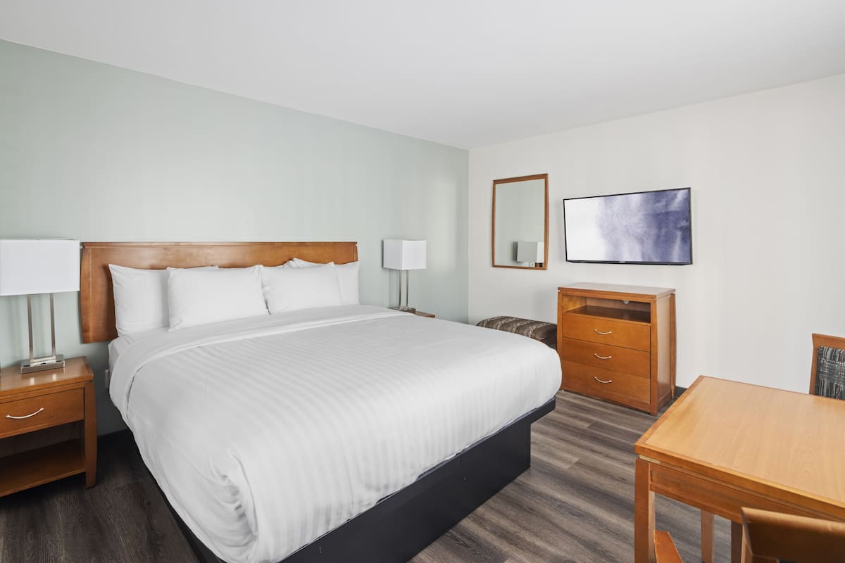 A hotel room with a large bed, a wooden nightstand with a lamp, a wooden dresser, a flat-screen TV mounted on the wall, and a wooden table with a chair.