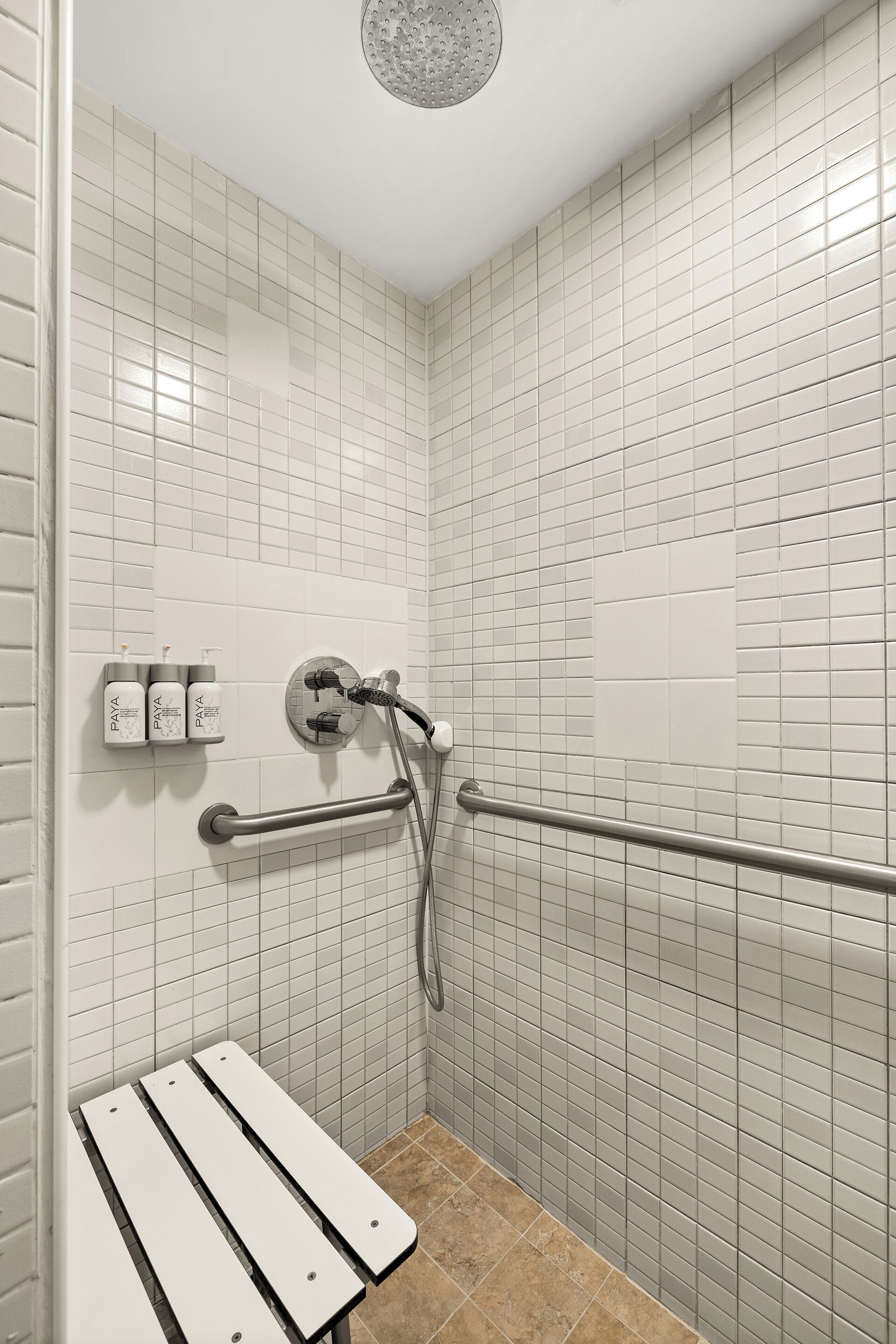 A shower area with white tiled walls, a handheld showerhead, a handheld shower attachment, and a bench.