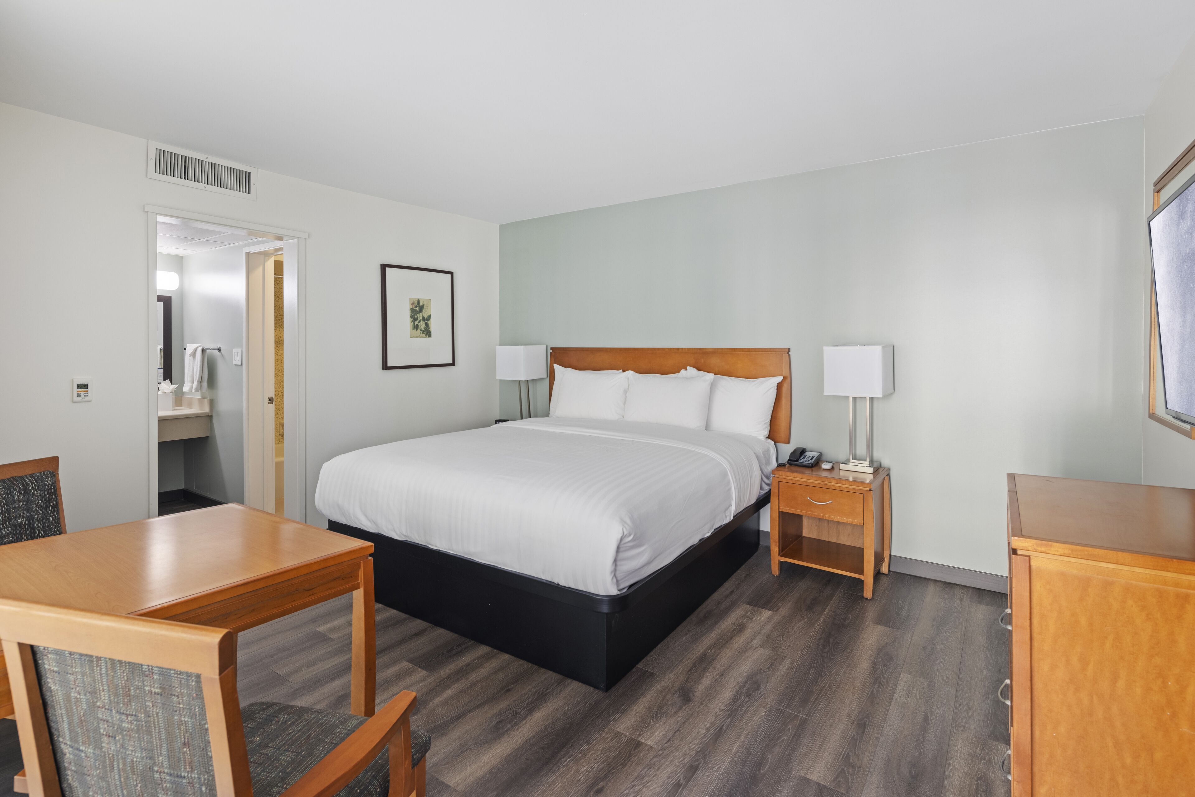 A hotel room with a large bed, a bedside table with a lamp, a wooden desk, and a chair.
