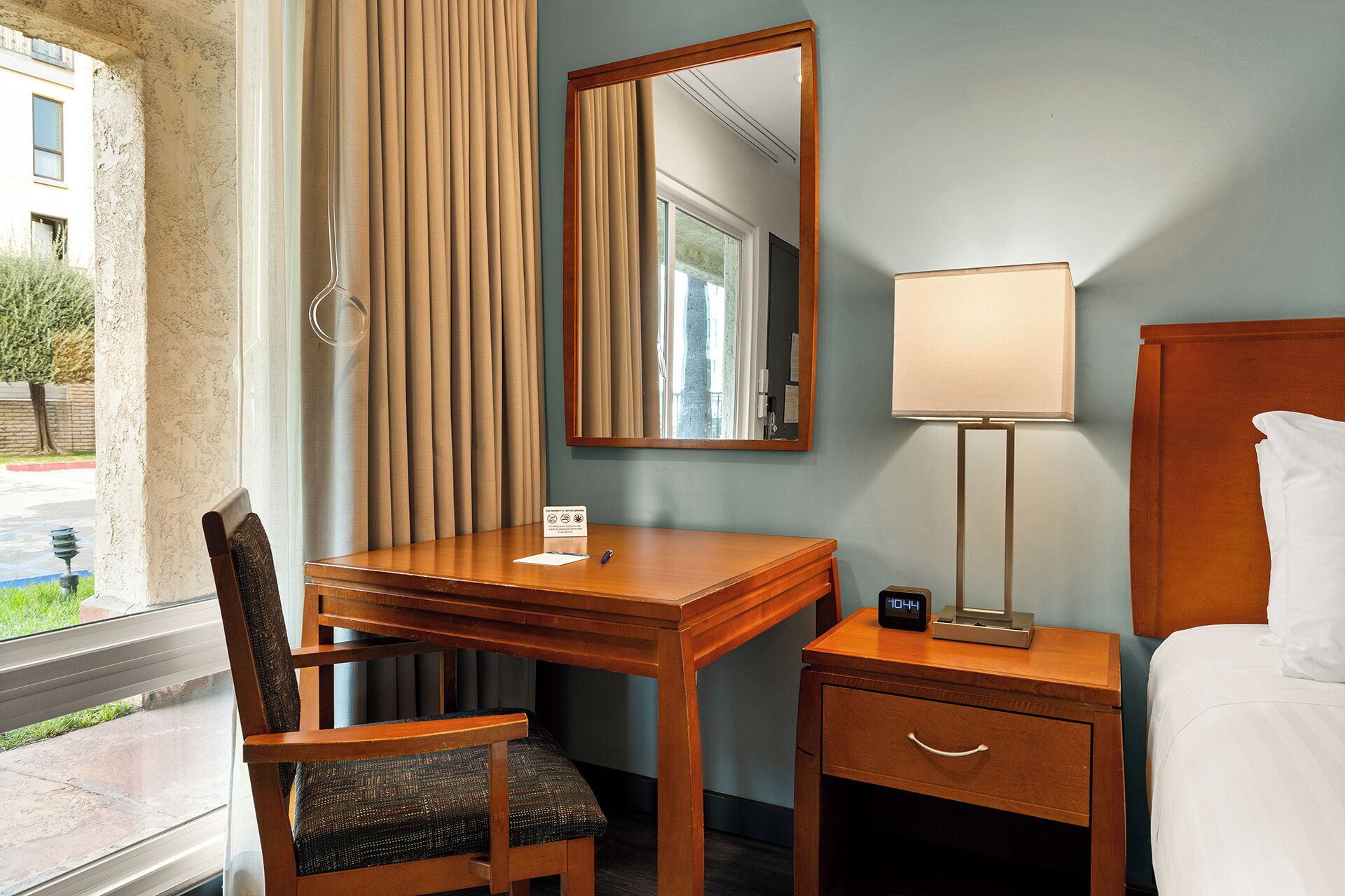 A hotel room with a wooden desk, a chair, a bedside table with a lamp, a bed, and a large mirror.