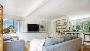 Ett modernt vardagsrum med en grå soffa, en platt-TV och en köksdel med vita skåp och ett matbord.