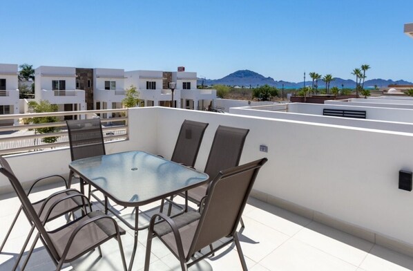 A rooftop terrace with a glass table and chairs, overlooking a coastal landscape with buildings and mountains.