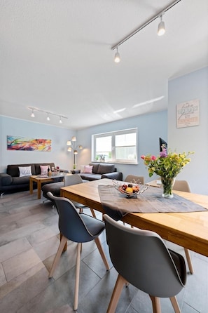 A modern living room with a wooden dining table, grey chairs, a sofa, and a large window. - Am Bahndamm 73 "Haus Meerblick" - bright and friendly vacation home in (Zingst)