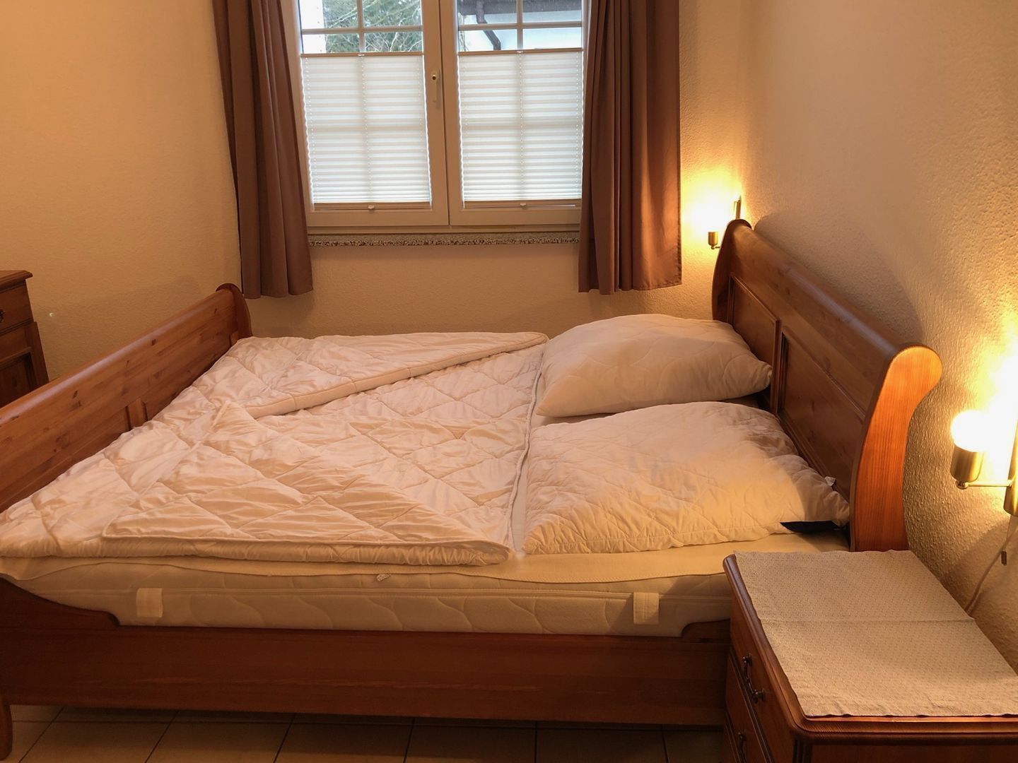 A neatly made bed with a wooden headboard, a bedside table with a lamp, and a window with curtains.