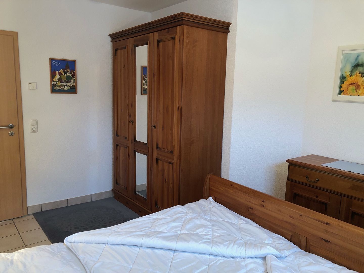 A bedroom with a wooden wardrobe, a bed with white bedding, a small dresser, and a painting on the wall.