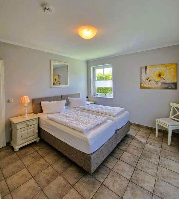 A double bed with a headboard, a bedside table with a lamp, a window with blinds, and a painting on the wall.
