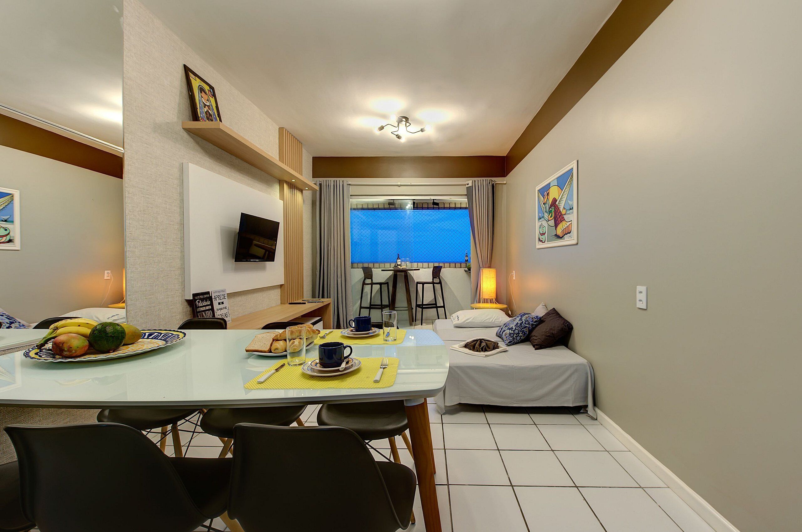 A dining area with a table set for two, a bed with pillows, a TV mounted on the wall, and a window with curtains.