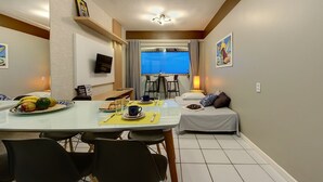 Un comedor con una mesa dispuesta para dos personas, una cama con almohadas, un televisor montado en la pared y una ventana con cortinas.