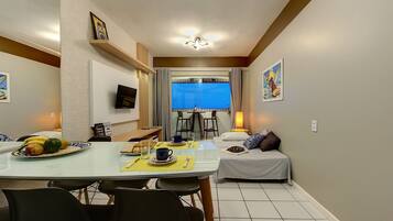 A dining area with a table set for two, a bed with pillows, a TV mounted on the wall, and a window with curtains.