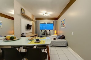 Uma sala de jantar com mesa posta para dois, cama com travesseiros, televisão na parede e janela com cortinas.