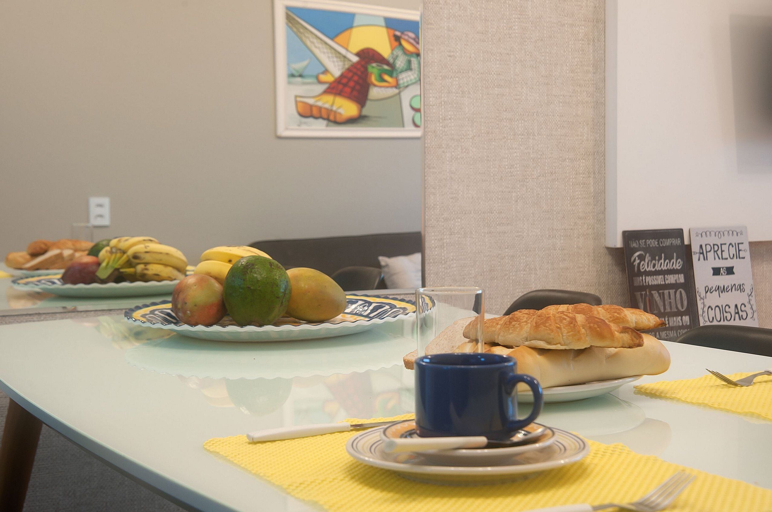 Disposição para o pequeno-almoço com pratos de fruta, um croissant e uma chávena azul sobre uma mesa com guardanapos amarelos.