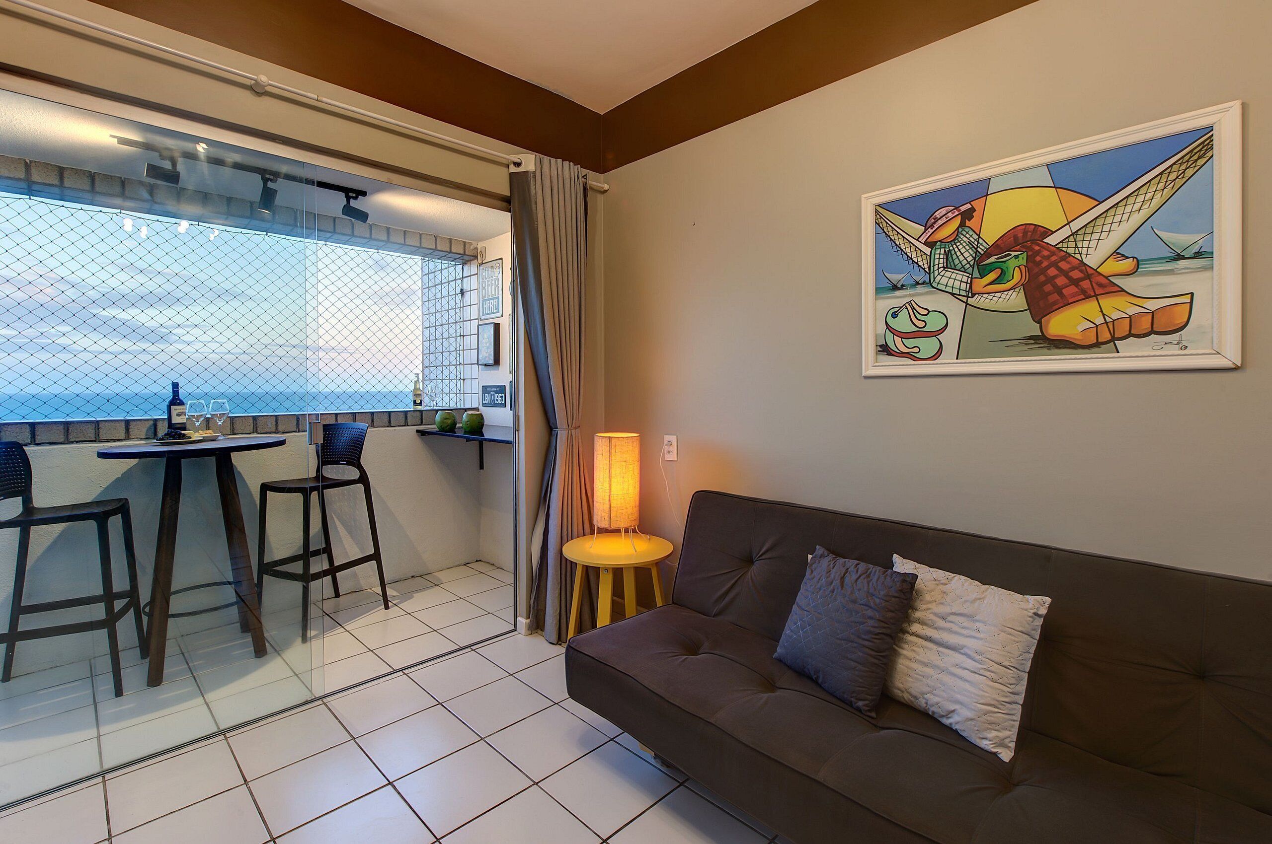 A living area with a sofa, a table, and two chairs. There is a window with a view of the sea and a painting on the wall.