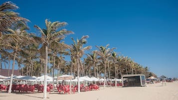Uma praia com fileiras de cadeiras vermelhas e guarda-sóis brancos, palmeiras e uma pequena construção na areia.