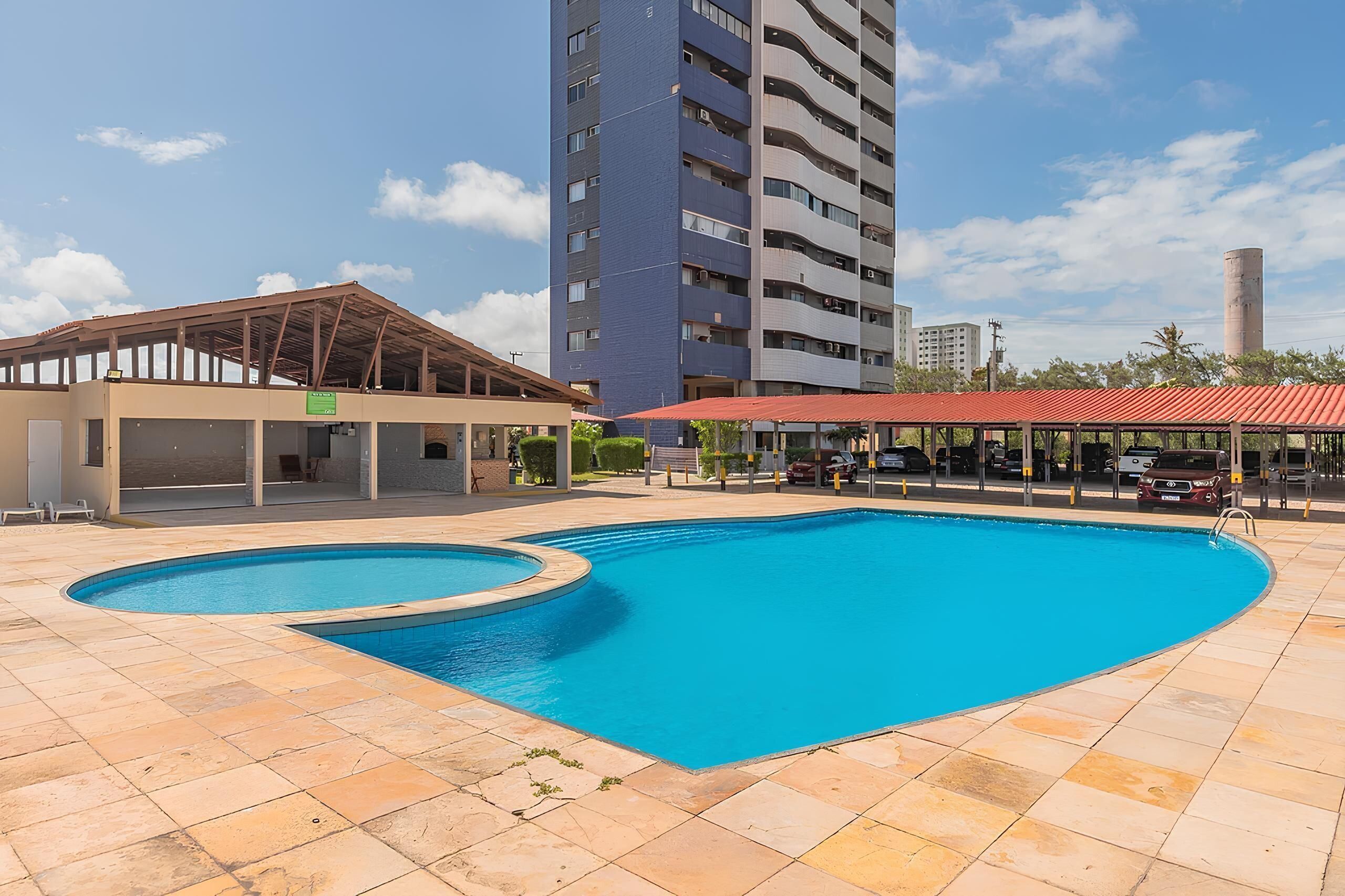 A swimming pool with a red-roofed pavilion and a covered area, surrounded by a high-rise building and a tall chimney.