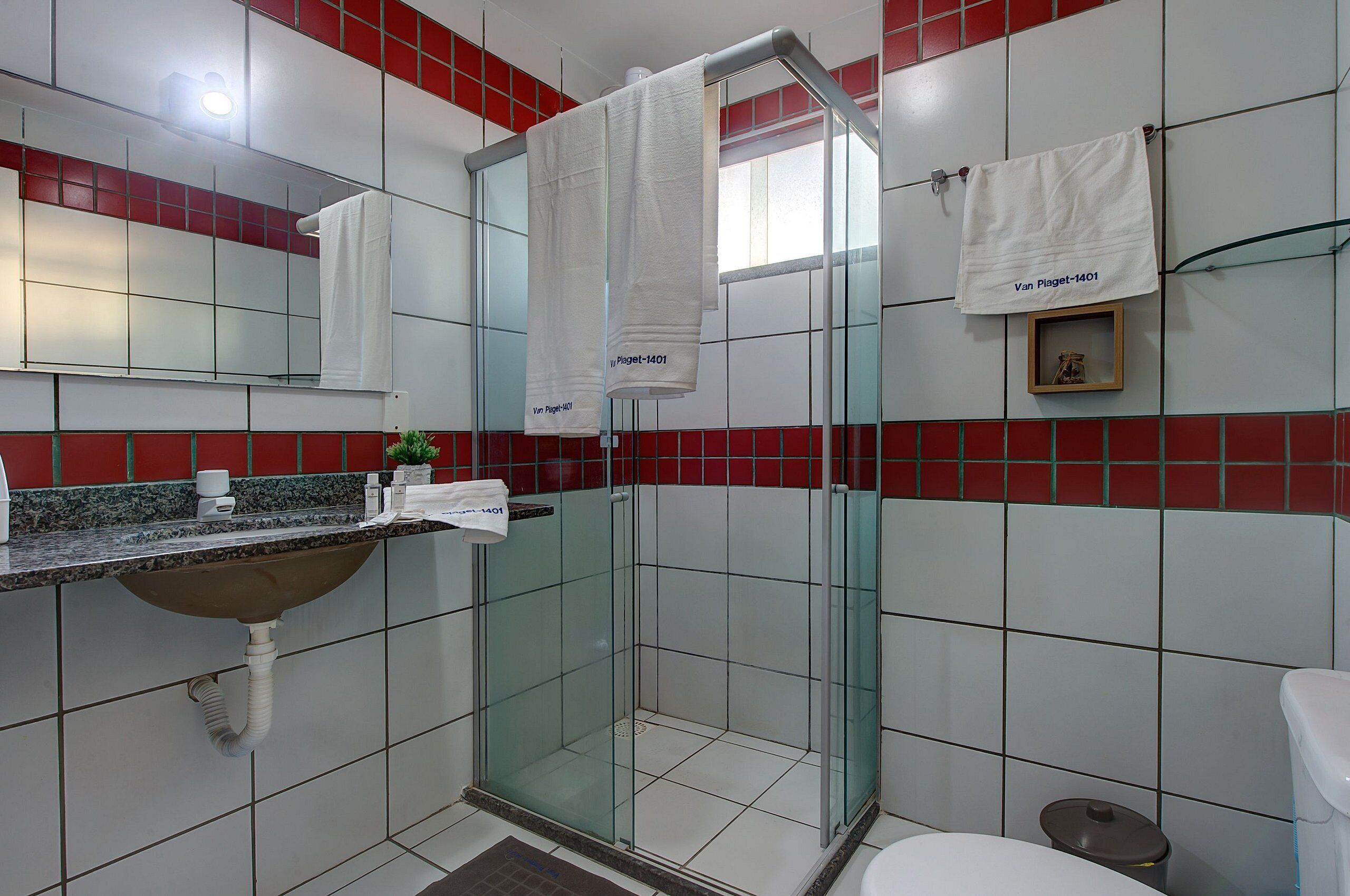 Un baño con ducha envuelta en vidrio, lavamanos con encimera de mármol y paredes con azulejos rojos y blancos.