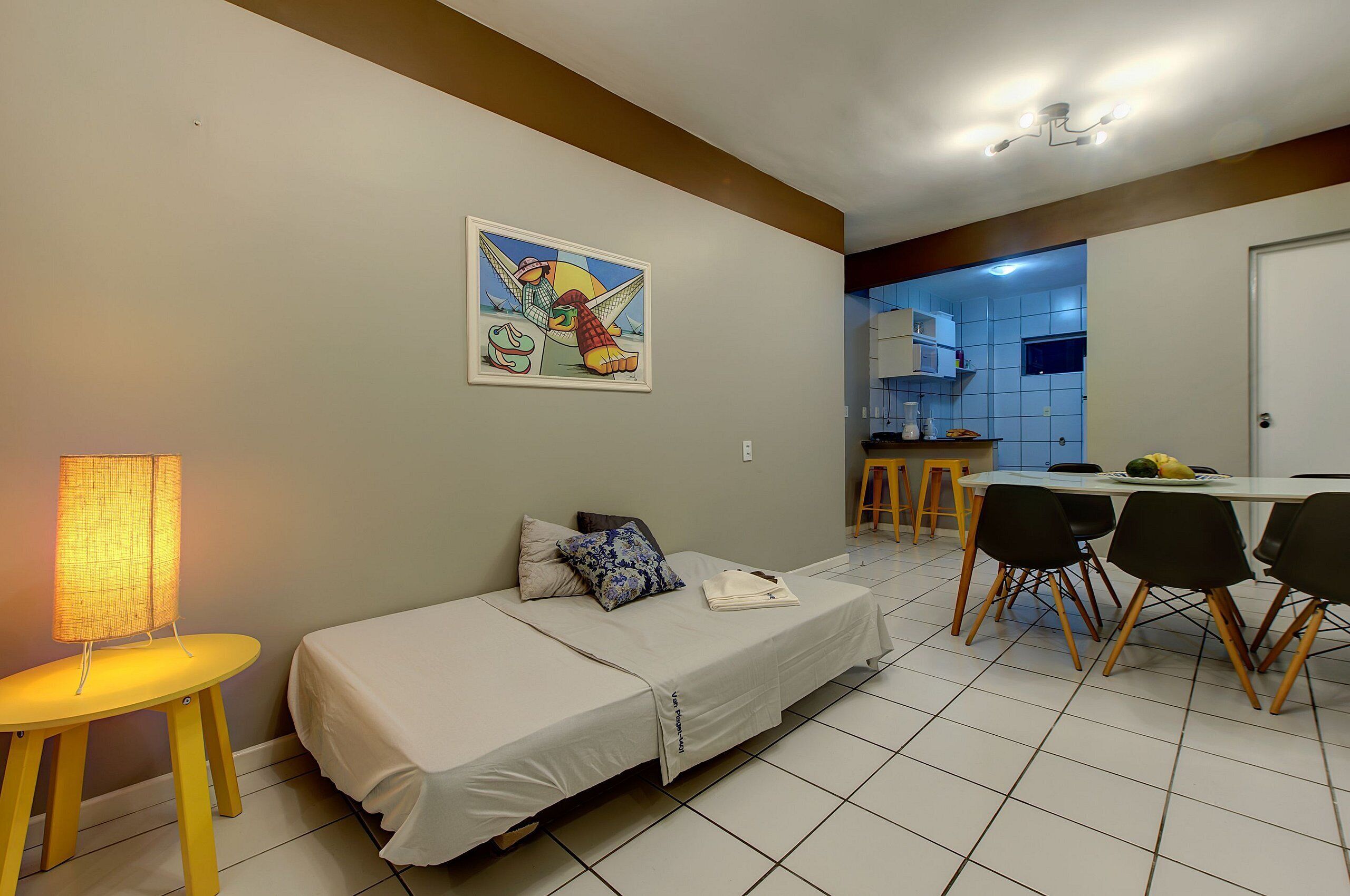 Une pièce de taille modeste comprenant un lit, une petite table avec une lampe, un coin repas avec des chaises et, au fond, une cuisine.