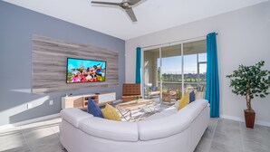 Una sala de estar moderna con un sofá blanco, un televisor de pantalla plana montado en una pared de paneles de madera, un ventilador de techo y un amplio ventanal corredera que da a un balcón con vistas a un parque y edificios a lo lejos.
