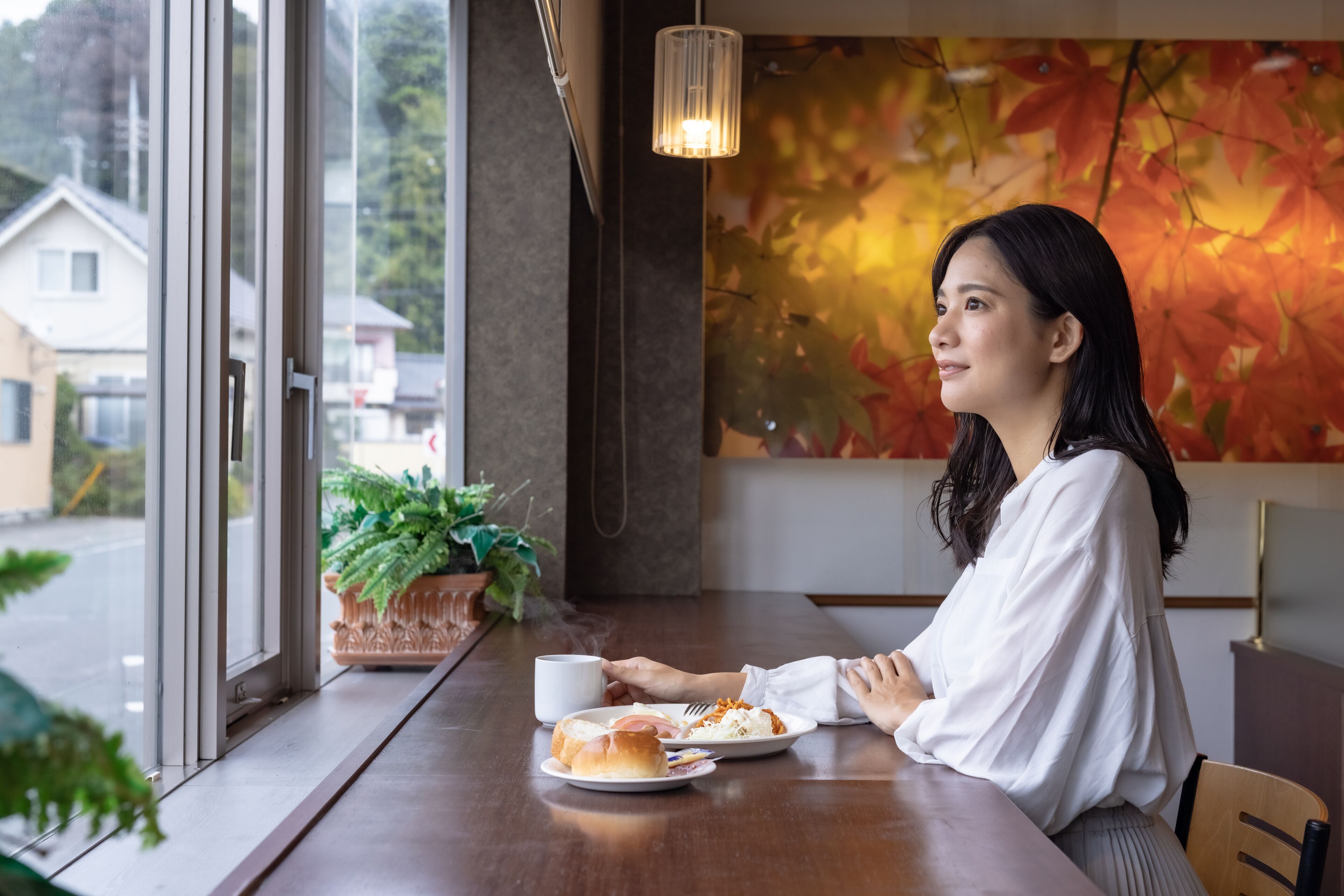 A person seated at a table with a cup and a plate of food, a plant on the windowsill, and a view of houses outside.