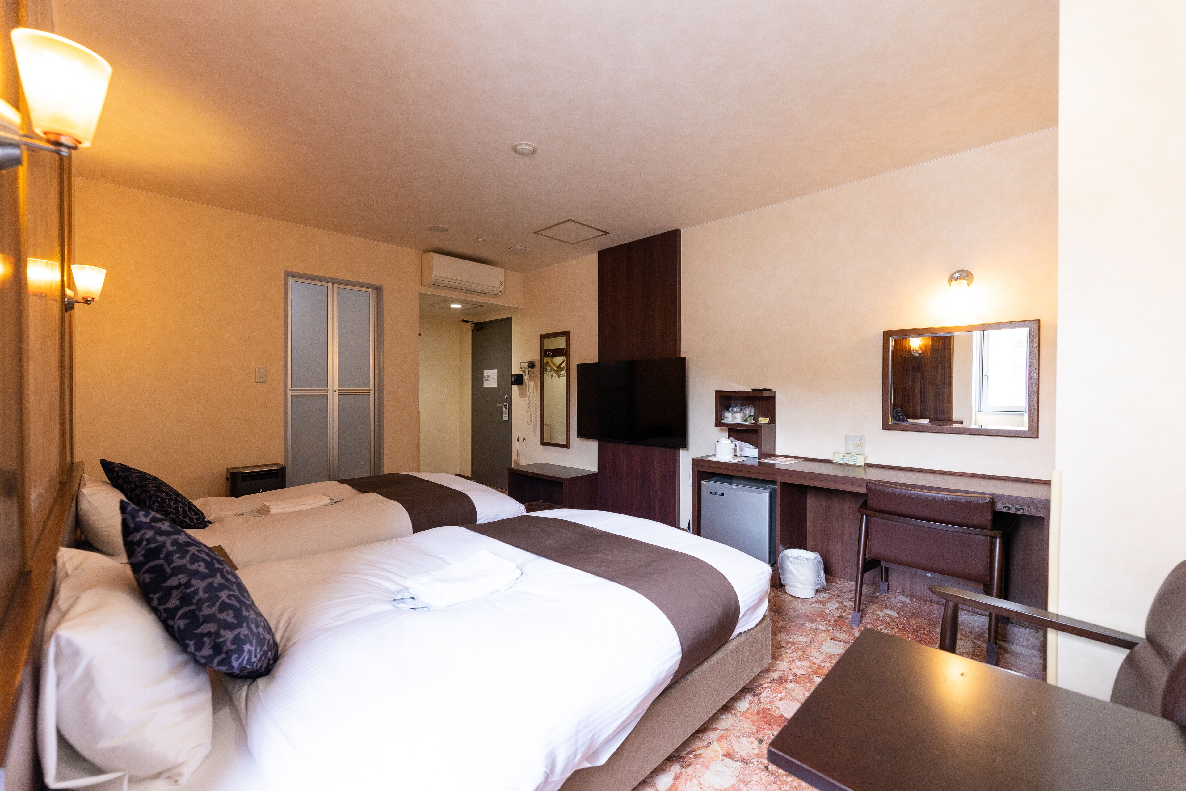A hotel room with two beds, a desk, a chair, a television, and a small kitchenette.