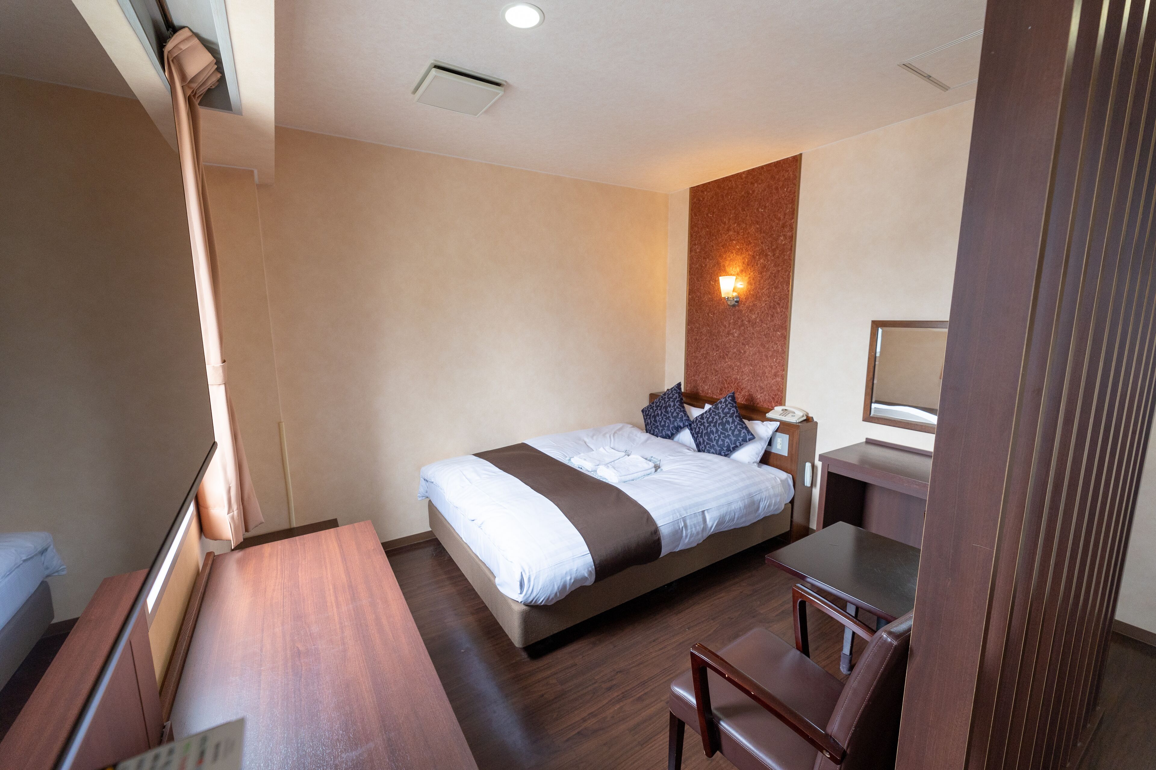 A hotel room with a bed, a desk, a chair, a bedside table, and a wall-mounted lamp.