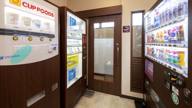 Ein Automatenbereich mit Cup Foods und Toyo Suisan Aufschriften, eine Tür mit Fenster und ein Schild, das einen Raucherbereich kennzeichnet.