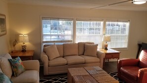 A living room with a beige sofa, a red chair, a woven ottoman, and two windows with blinds. - Beach Cottage - family and pet friendly, beach access less than 100ft away (Fernandina Beach)