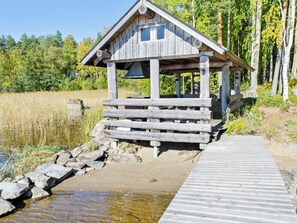 Una cabaña de madera con un porche, un muelle de madera y un paisaje natural con árboles y una masa de agua.