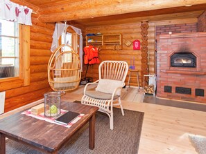 A cozy log cabin interior with a wooden table, wicker chairs, a fireplace, and a window with curtains. - Suvituuli by Interhome (Liperi)