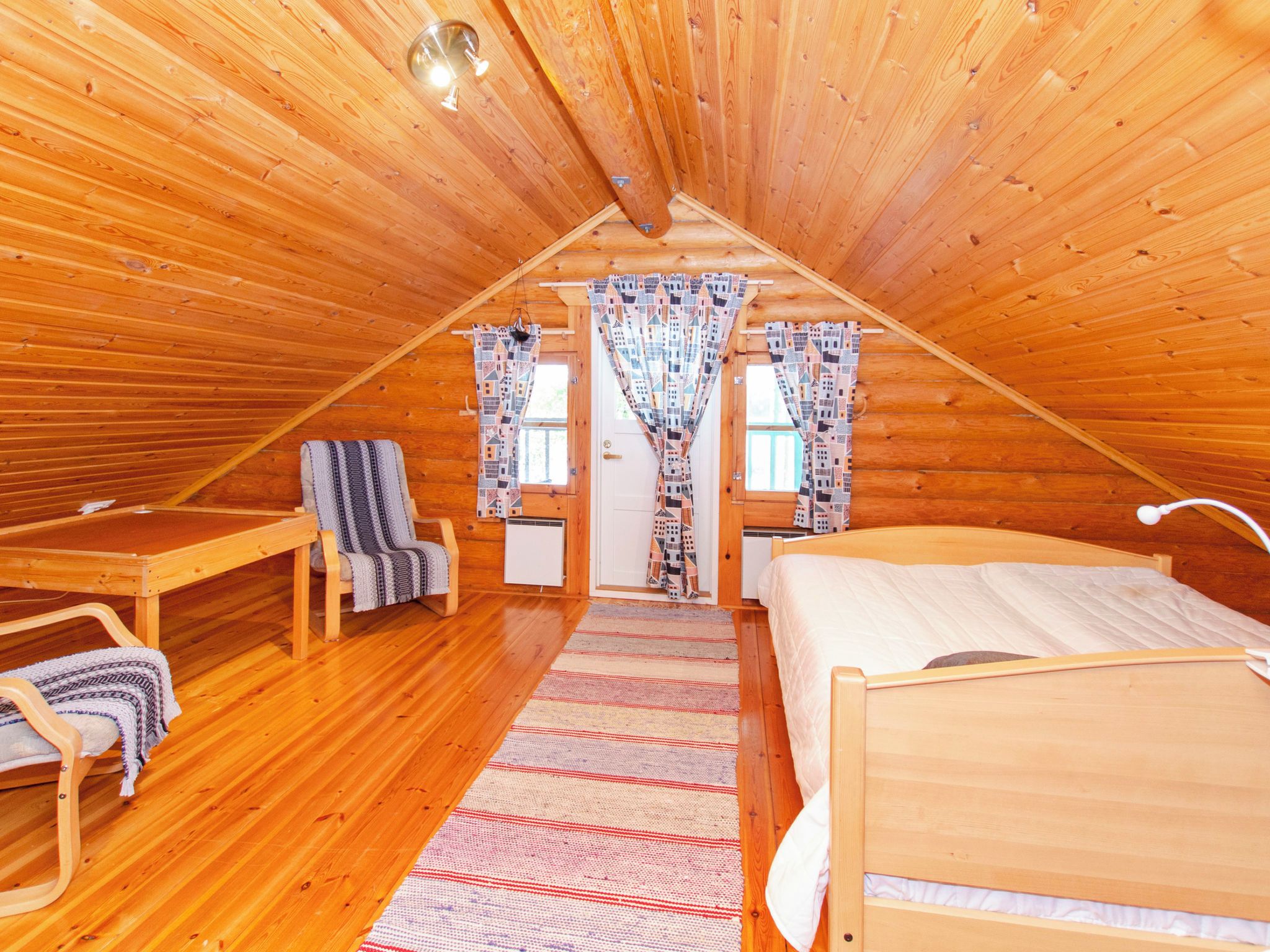 A cozy attic bedroom with a wooden sloped ceiling, a single bed, a chair, a small table, and a window with patterned curtains.