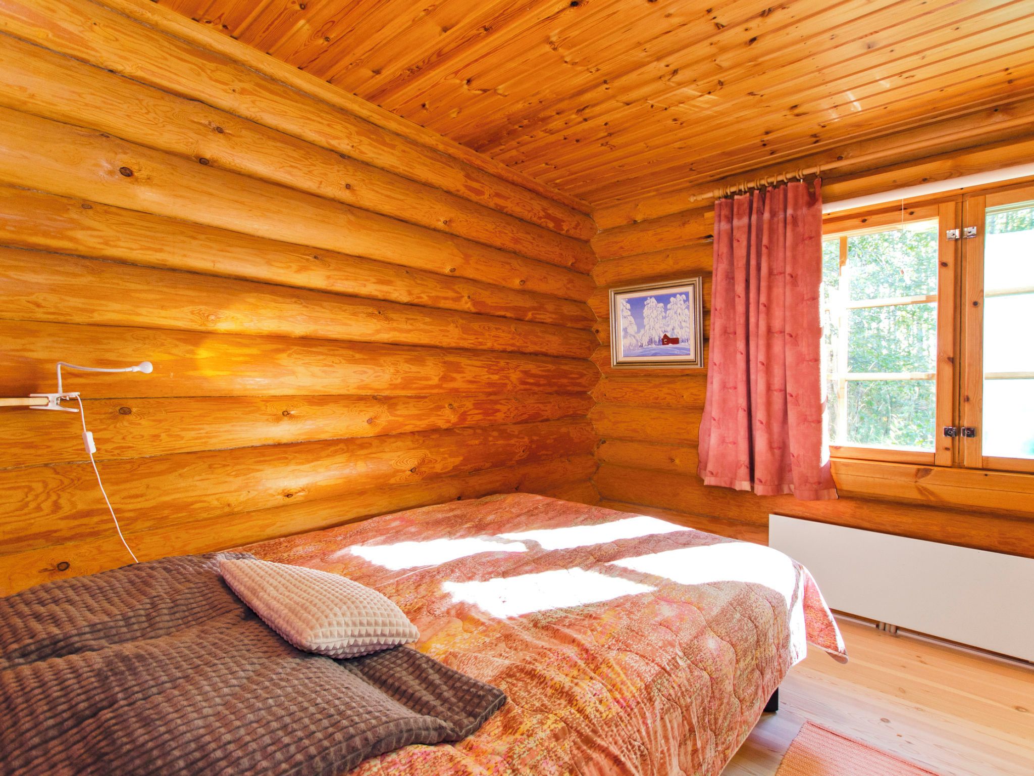 Ein Zimmer in einer Holzhütte mit einem Bett, einem Fenster mit Vorhängen und einem Bild an der Wand.