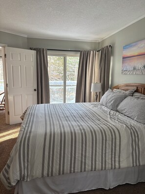 A bedroom with a bed, a nightstand, a lamp, a door, and a window with curtains. - Cozy and Comfy 2 bedroom condo (Bellaire)