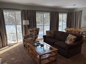 A living room with a sofa, armchair, and coffee table, large windows with curtains, and a view of a snowy outdoor area. - Cozy and Comfy 2 bedroom condo (Bellaire)