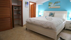 A bedroom with a bed, a wooden wardrobe, a nightstand, and a wall with a tropical painting. - Tranquil Oceanside: Pools & Direct Beach Access (Sint Maarten)