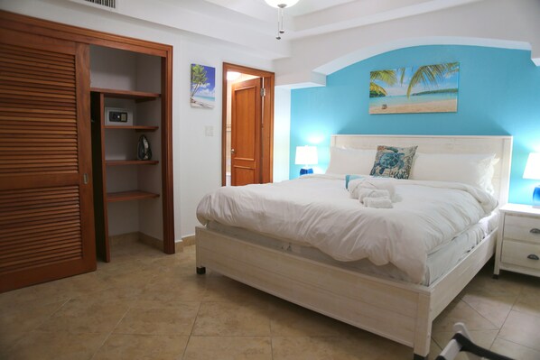 A bedroom with a bed, a wooden wardrobe, a nightstand, and a wall with a tropical painting.