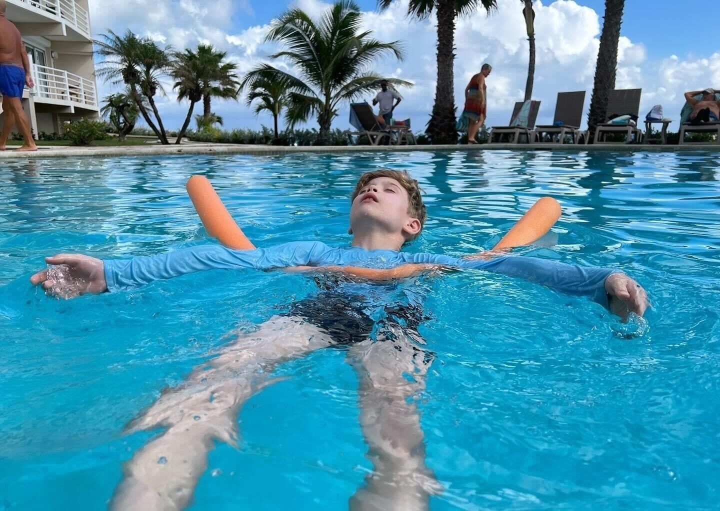 A person floating in a pool with a clear sky and palm trees in the background.