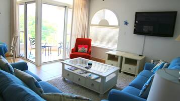 Un salon comprenant un canapé bleu, un fauteuil rouge, une table basse en verre et un téléviseur à écran plat fixé au mur.