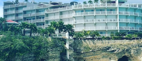 A multi-story hotel with balconies, located near a beach with clear turquoise water.