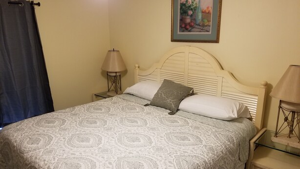 A neatly made bed with patterned bedding, two bedside lamps, a nightstand with a glass top, and a framed picture on the wall. - Beach-living 2 bed (Fernandina Beach)