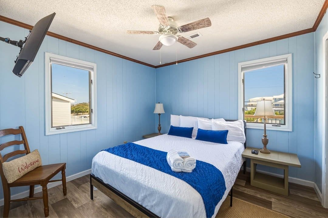 A bedroom with a bed, a ceiling fan, two windows, a chair, and a nightstand.