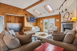 A cozy living room with a fireplace, wooden walls, and a large window. - Sandpiper Paradise | 630 ft to the beach | Private Pool (Virginia Beach)