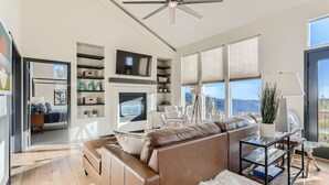 A spacious living area with a large brown leather sofa, a glass coffee table, and a ceiling fan. There is a television mounted on the wall, and a balcony with a view of the outdoors. - Cozy Mountain Escape | Hot Tub +Views | Dog Friendly | Sleeps 10 | 45 min Denver (Evergreen)