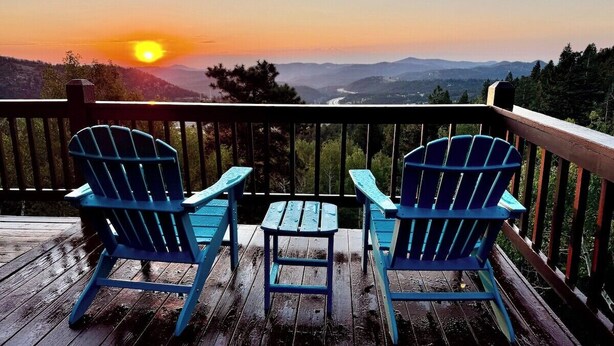 A wooden deck with two blue Adirondack chairs and a small table, overlooking a scenic view of mountains and a sunset. - Cozy Mountain Escape | Hot Tub +Views | Dog Friendly | Sleeps 10 | 45 min Denver (Evergreen)