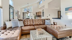 A spacious living room with a large brown leather sectional sofa, a glass coffee table, and a dining area with a chandelier. - Cozy Mountain Escape | Hot Tub +Views | Dog Friendly | Sleeps 10 | 45 min Denver (Evergreen)